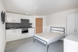 A studio room in 59 West Street. Against the far wall is a kitchen unit with an oven/hob, sink, fridge/freezer and cupboard space. To the right of the kitchen unit is the door to the room. A double bed is the right of the door and a television is mounted onto the wall opposite the bed. To the right of the bed is a wardrobe and chest of drawers.